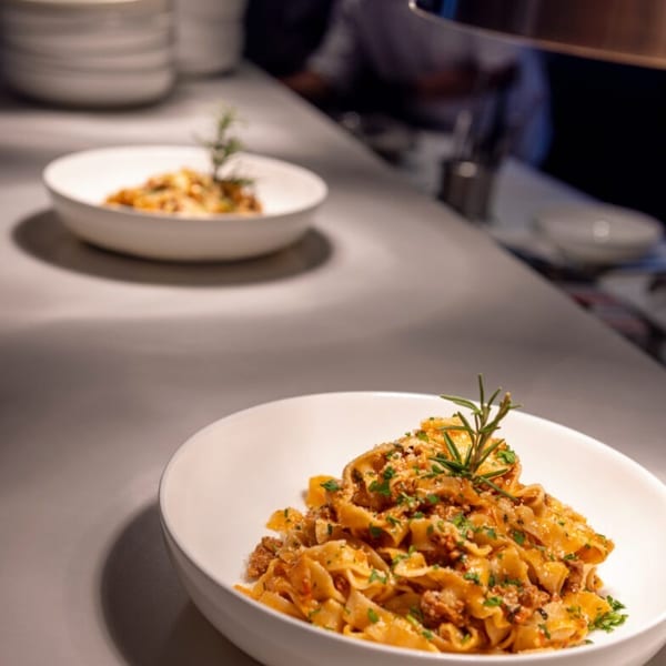 Two bowls of pasta with meat sauce and rosemary on a kitchen counter, with a chef in the background.