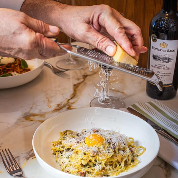 Hands grating cheese over a bowl of pasta carbonara with an egg yolk, next to a bottle of red wine.