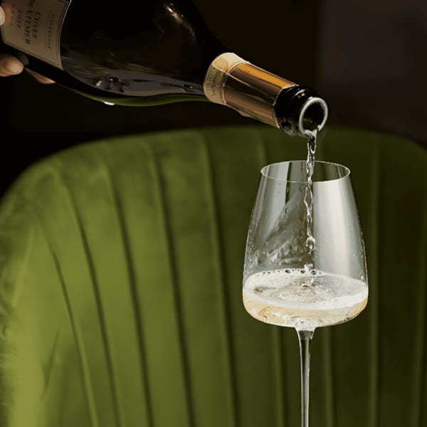 Sparkling wine being poured from a bottle into a glass, with a green velvet chair in the background.
