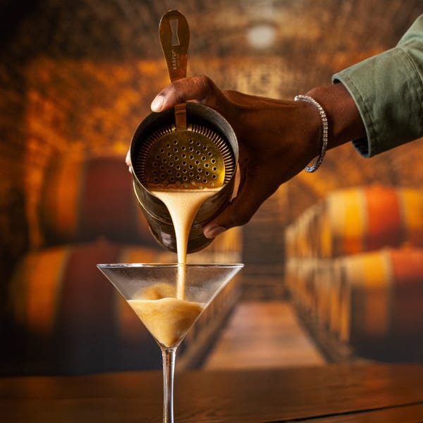Bartender pouring a creamy cocktail from a shaker into a martini glass on a wooden bar.