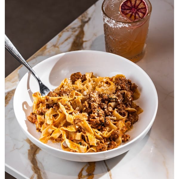 Fettuccine bolognese with parmesan cheese and a cocktail with a blood orange slice on a marble table.